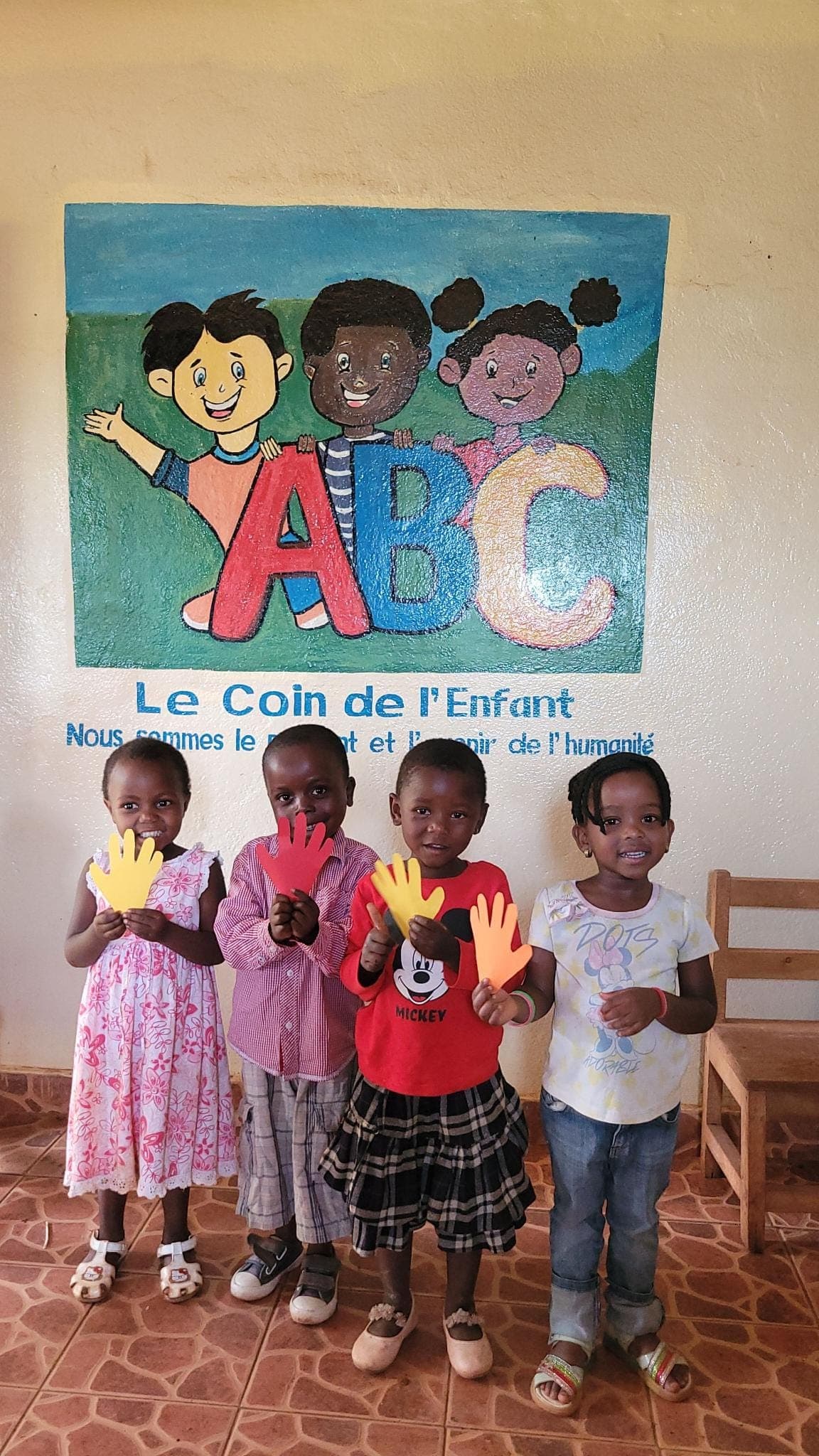 Kinder halten bunte Papierhände vor dem Wandbild „Le Coin de l’Enfant“ — Kinderprojekt im Ausland, unterstützt von der Kette der Solidarität e.V.