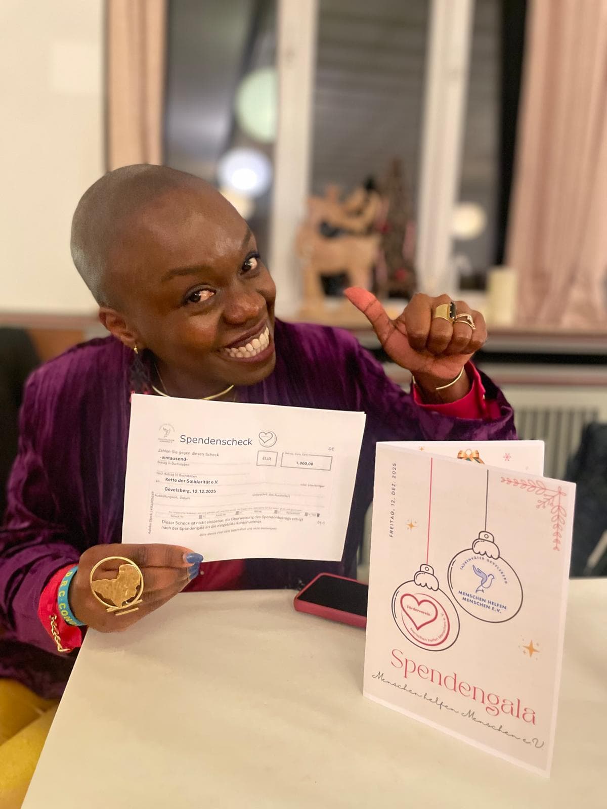 Yasmine Bisimwa von der Kette der Solidarität e.V. hält lachend den Spendencheck über 1.000 € und die Programmkarte der Spendengala „Menschen helfen Menschen e.V.“ vom 12. Dezember 2025